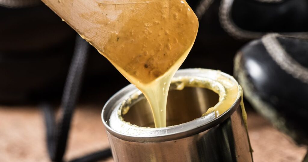 Yellow coloured adhesive glue liquid dripping from a wooden spoon into a silver tin.