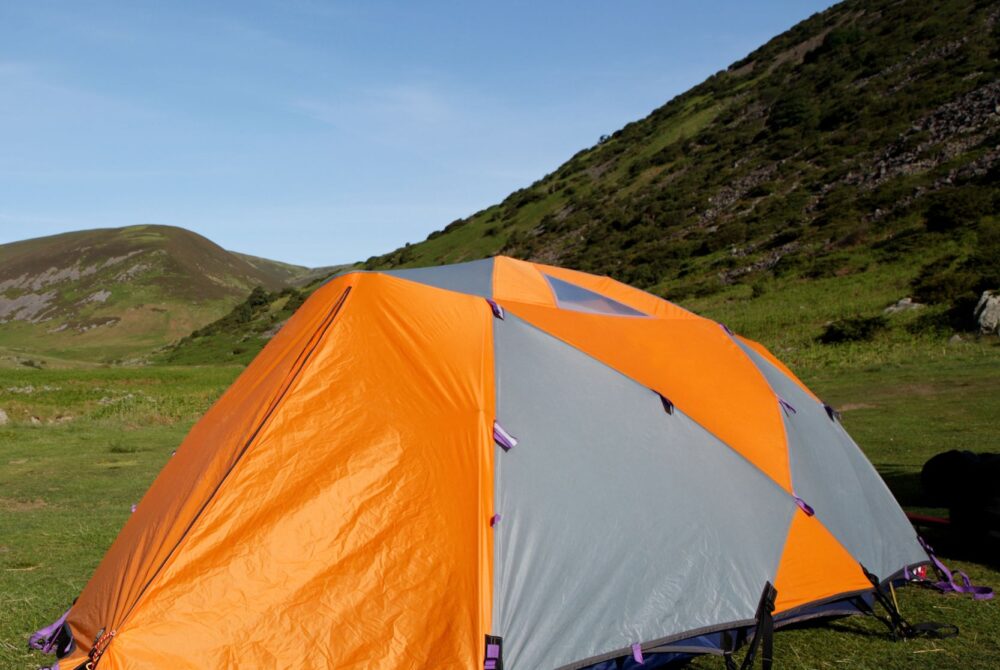 Small orange and grey tent pitched on a grass hill side blue skies.