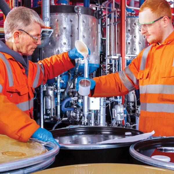 Two male factory process operators in orange overall decanting coating material.