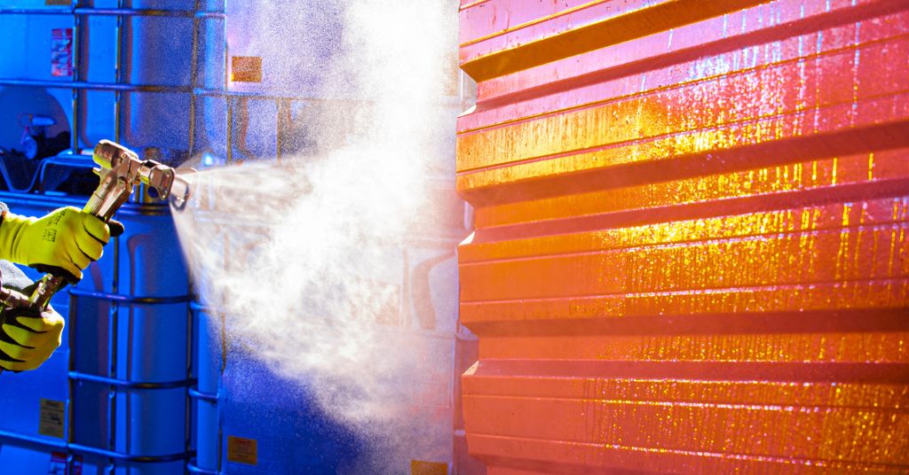 Person in yellow safety gloves conducting spray coating applications on some orange metal cladding with a paint spray gun.